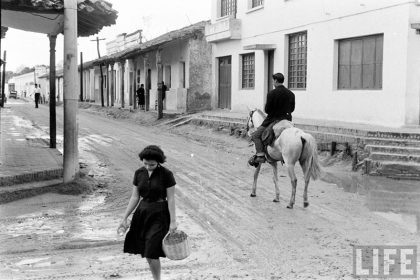         Mujer cruzando la calle con una canasta y detrás de ella una persona a caballo sobre la calle Independencia de ida a la calle Mercado
