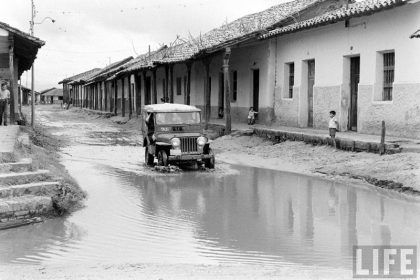         Taxi Jeep Willys con pasajeros cruzando el charco de agua formado en la calle Pari esquina Independencia – fotografía 1 de 2
